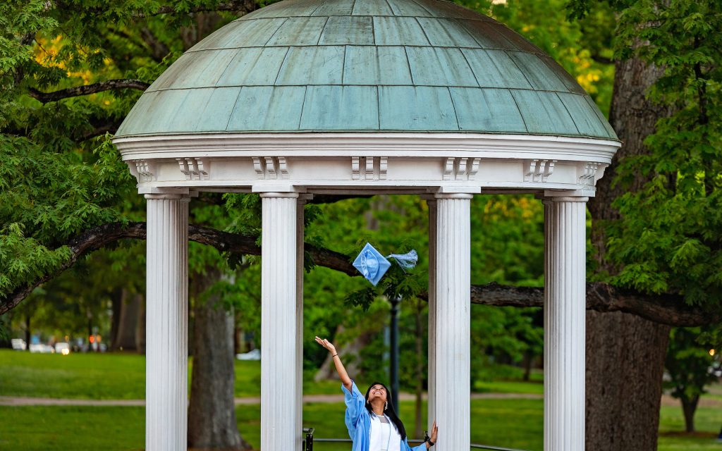 A girl in cap and gown tosses her Commencement cap in the air in front of the Old Well.