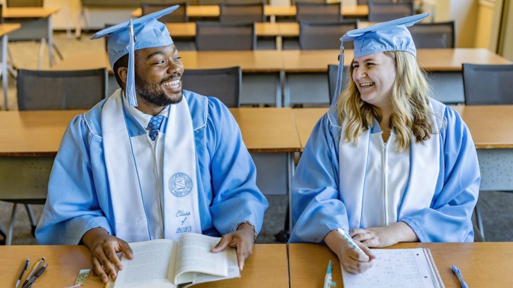 DeAndre Sawyer and Kacie Horton sit at desks in a classroom smiling at each other, wearing their caps and gowns.