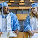 DeAndre Sawyer and Kacie Horton sit at desks in a classroom smiling at each other, wearing their caps and gowns.