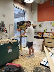 Cy stands surrounded by wood tools and other BeAM makerspace tools as he smiles at the camera.