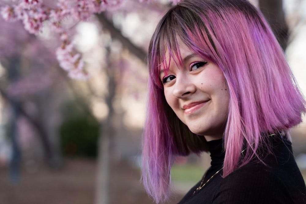 A headshot of Victoria Wlosok, who has short, pink hair. Behind her, cherry blossoms bloom.