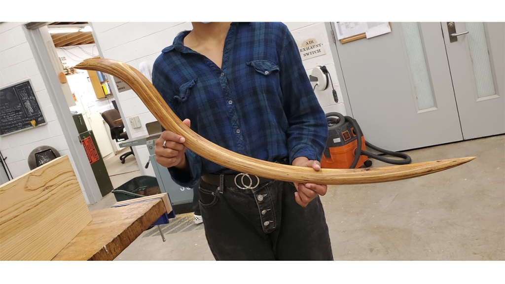 A student holds a long, thin wood sculpture that resembles an archer's bow.