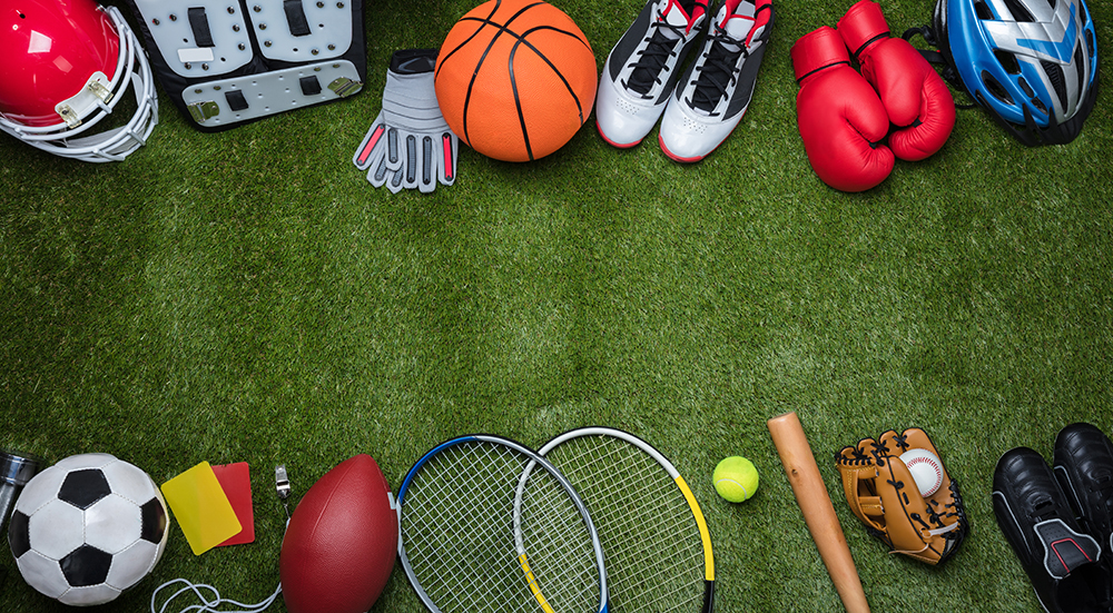 A row of sports equipment for various sports lies on a grassy field. Examples include a basketball, tennis rackets, sport shoes, a baseball bat, etc.