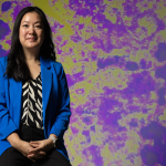 Professional waist-up shot of Angel Hsu in front of a mottled green and purple background.