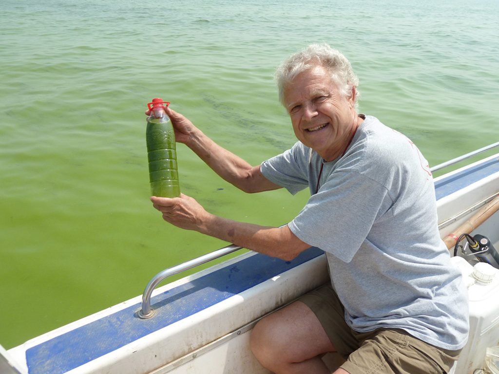 Hans Paerl sits in a bot holding a water bottle full of green algae-colored water.