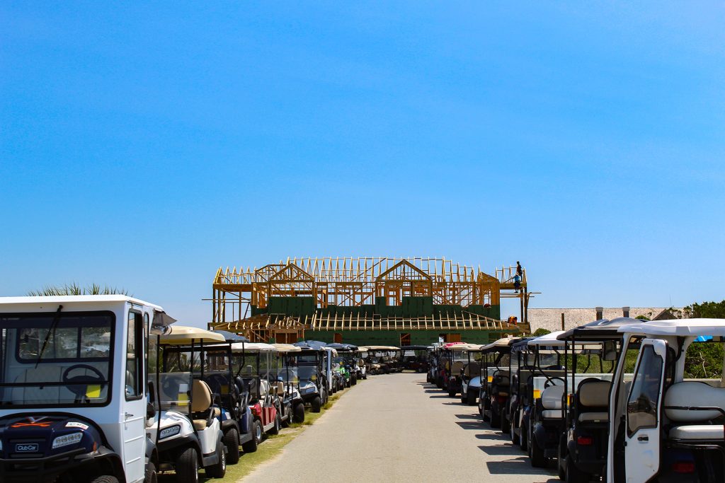 A row of golf carts is lined up in front of a public beach access point where a large new home is under construction.