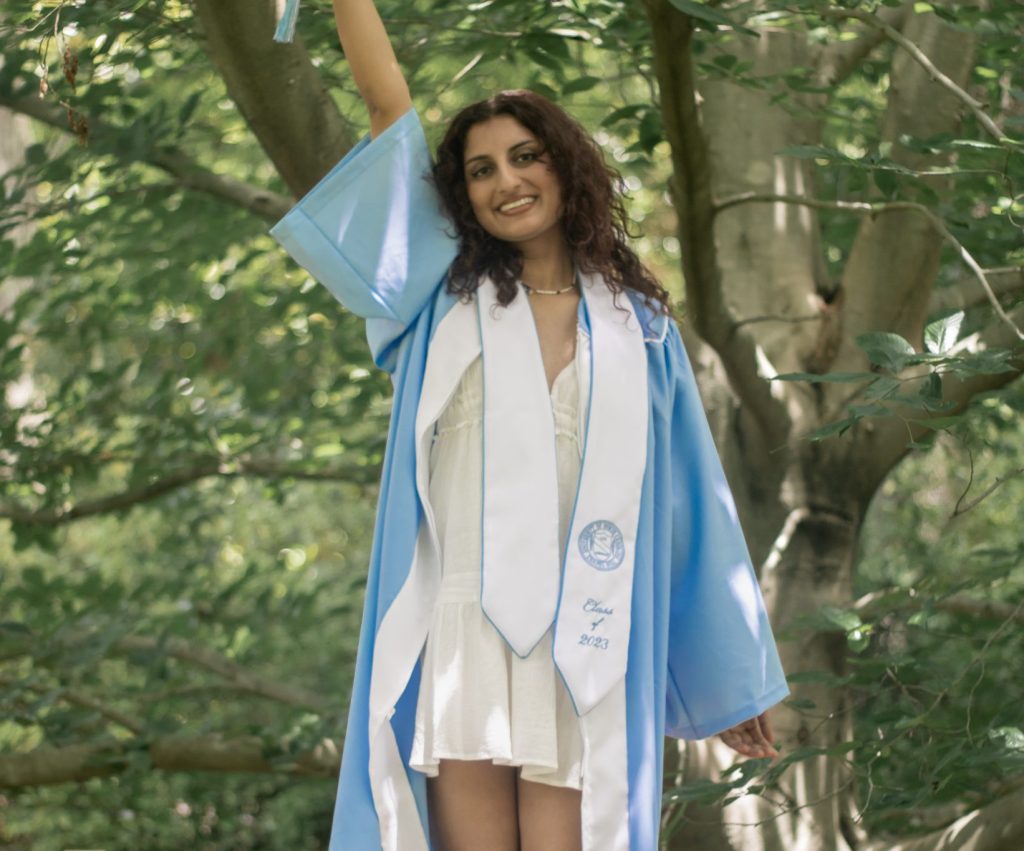 Avni wears her cap and gown and stands on a bench in the arboretum