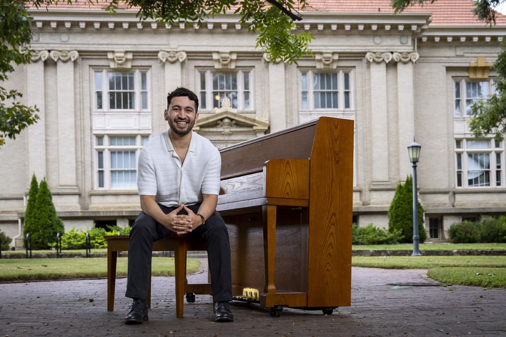 Hector Aizpurúa poses on a piano bench, facing away from the piano. Hill Hall, the music building, is in the background.