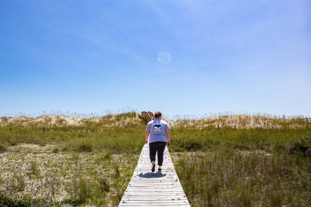 The back of Kimmy Hansen as she walks down a plankway toward the beach.
