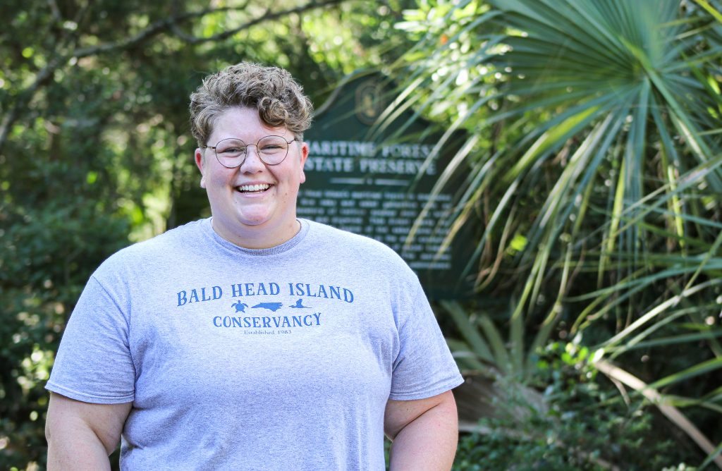 Headshot of Kimmy Hansen, who wears a Bald Head Island Conservancy shirt. She stands in front of trees, which make up the maritime forest she researches.