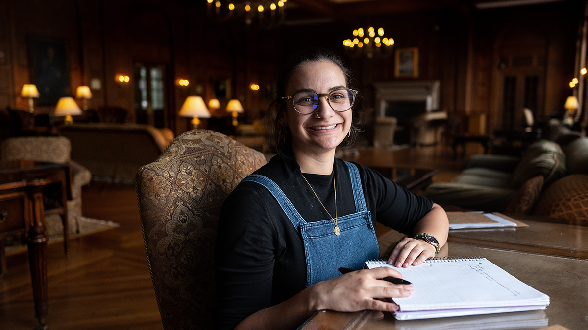 Headshot of Sara Rodrigues sitting with her notebook at a table in the Graham Memorial Lounge.