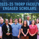 A group photo of 15 faculty members standing together and posing for a photo outside in front of a brick wall with trees behind them.