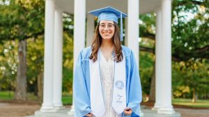 Headshot of Emma Cohn in her Carolina blue graduation cap and gown, the Old Well in the background.