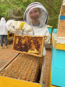Jordan wears a white beekeeping suit and holds a bee hive.