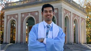 Headshot of Pramit Thapa in a Carolina blue graduation gown, the Bell Tower in the background.