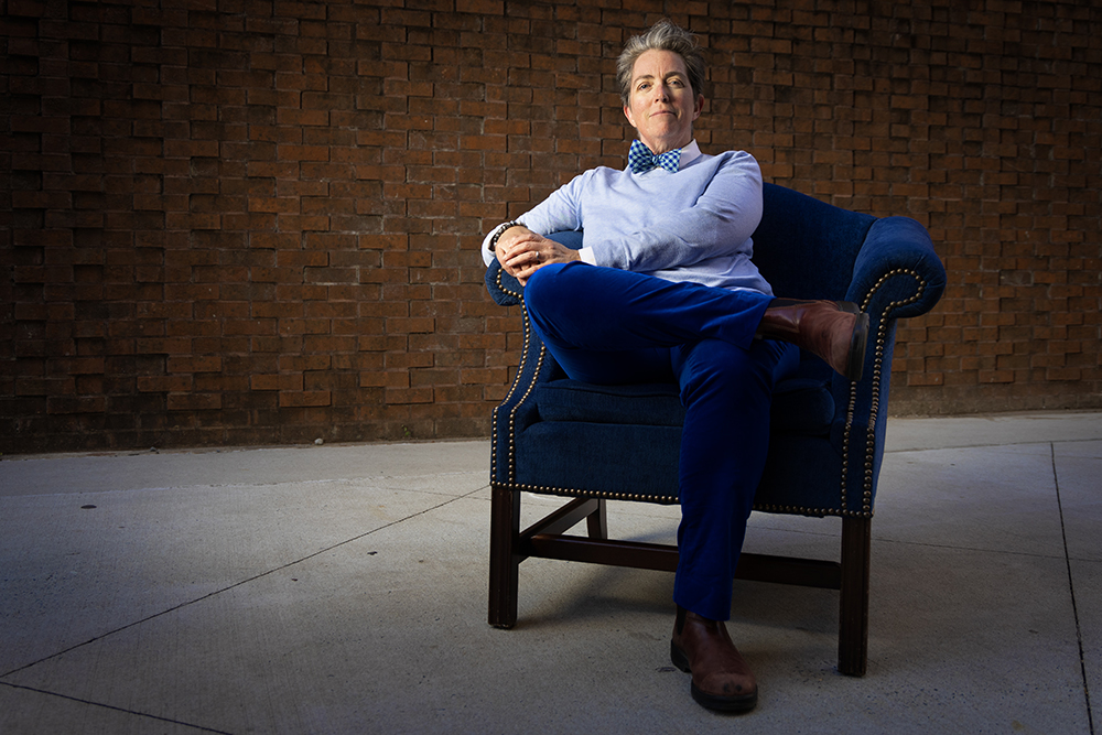 Gabrielle Calvocoressi sitting in a plush blue chair in front of a stark brick background.