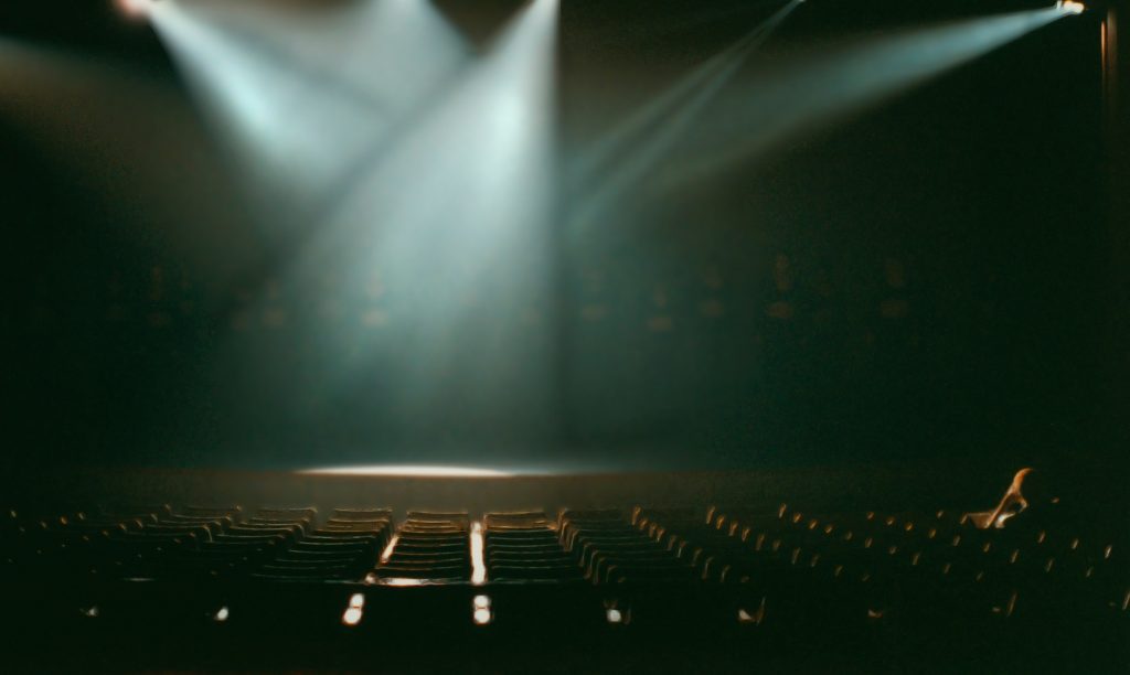 Stock photo of the interior of a theater, with several overhead lights illuminating an empty stage.