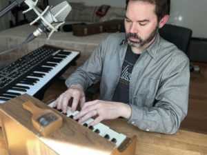 Lee Weisert plays a toy piano at home, where a full keyboard and microphone is set up to the side and his bed is behind him.