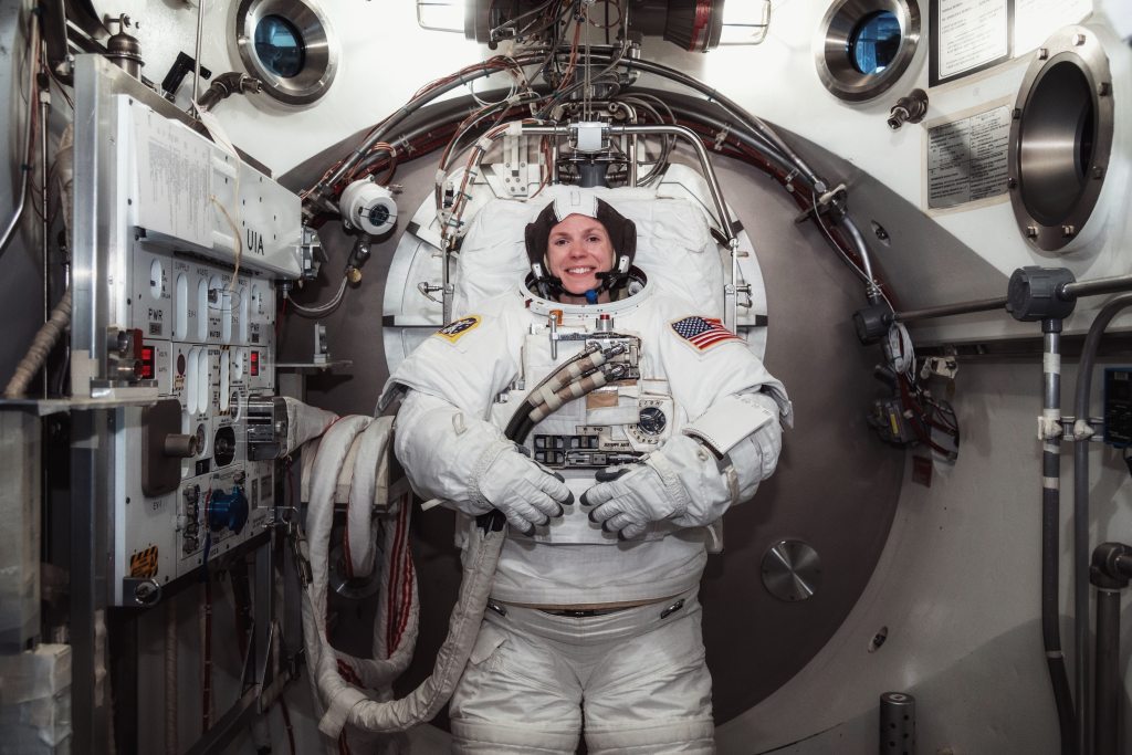 Zena Cardman stands and smiles at the camera in her astronaut gear, surrounded by space equipment.