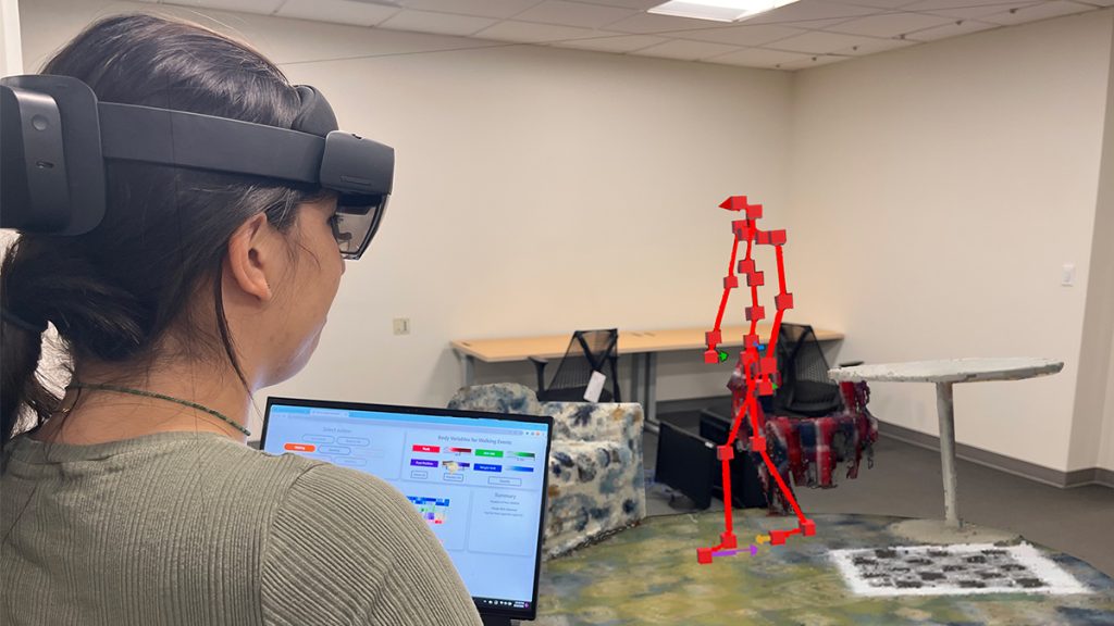 A researcher holding a laptop and wearing augmented reality glasses examines a model of a person, a red stick figure-like construction, through the AR technology.