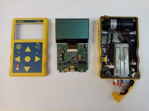 Three components of a deconstructed yellow Geiger counter: (l-r) the basic keyboard, the circuit board attached to the screen, and the battery compartment. 