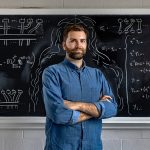 Andrew Adair stands in front of a chalkboard with mathematical formulas written on it.