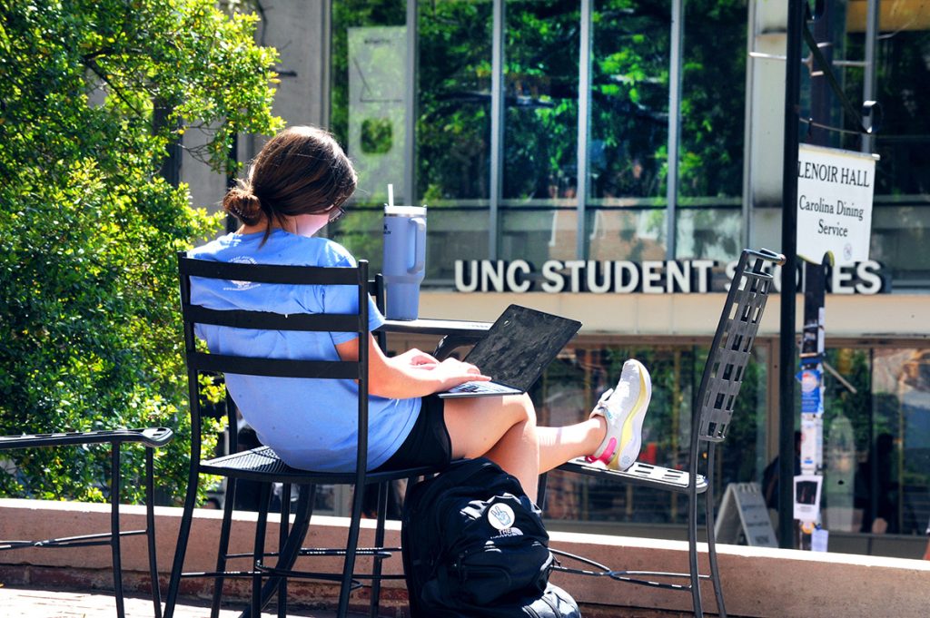 A student sits on a bench outside student stores and works on a laptop. You can see the Student Stores sign across from the student.