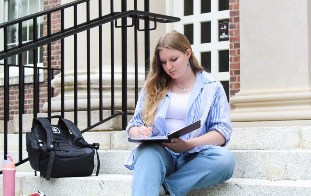 Emma Gates sits on the big stone steps outside Manning Hall, writing in a notebook.