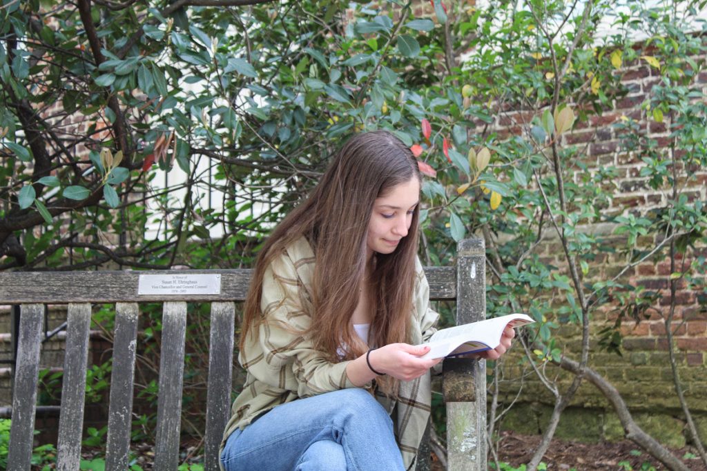 Frances Norton reads a book as she sits on a bench, greenery behind her.