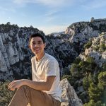 Austin Vo sits on a mountain top. Behind him are mountains and cliffs under a blue sky.