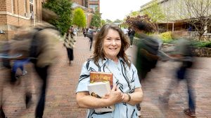 Janet Vitolo stand in the middle of campus holding books while blurred images of fellow students walking surround her.