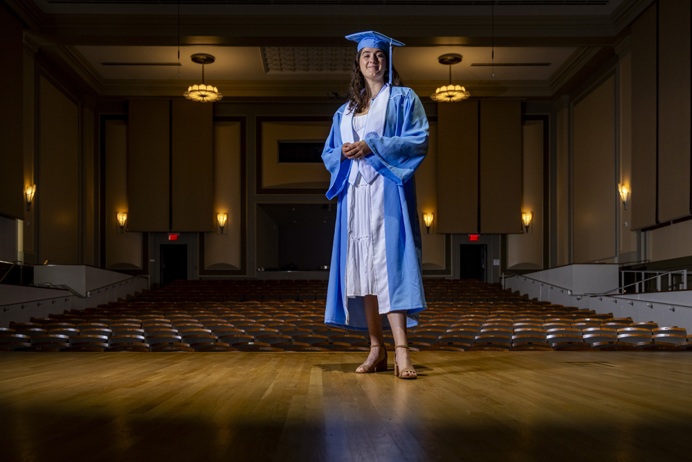 Carrina Macaluso is a graduating senior majoring in music. Sh stands wearing cap and gown on the stage in Hill Hall. 