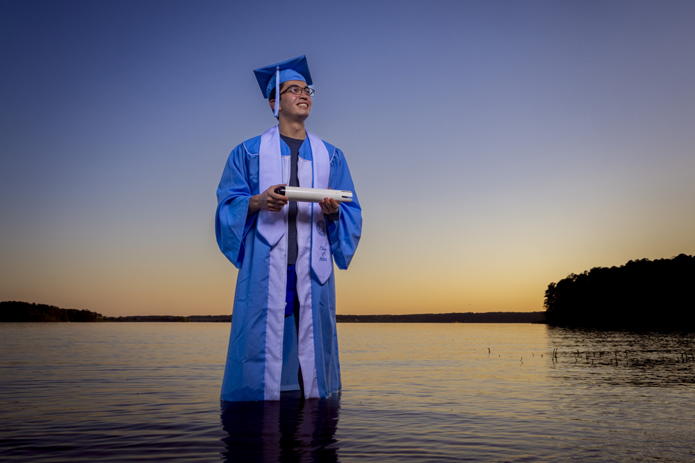 David Go is a graduating senior majoring in Geological Sciences within the UNC College of Arts and Sciences. He stands in the middle of a lake wearing his cap and gown. 