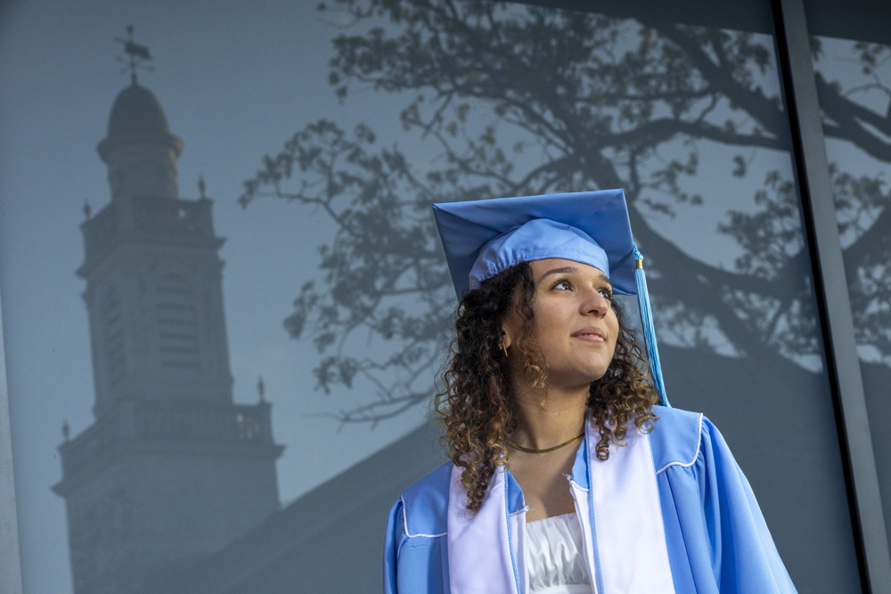Malak Dridi is a graduating senior -- she stands in front of a window with the Carolina campus in the background. She is wearing a cap and gown.