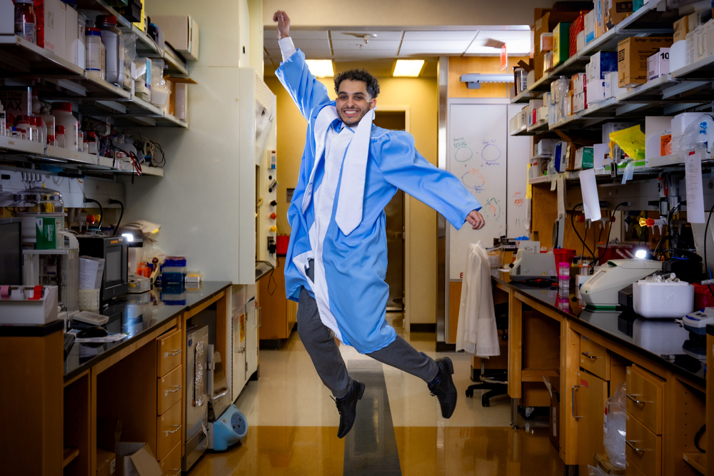 Rami Darawsheh jumps in the air wearing a cap and gown in a lab.