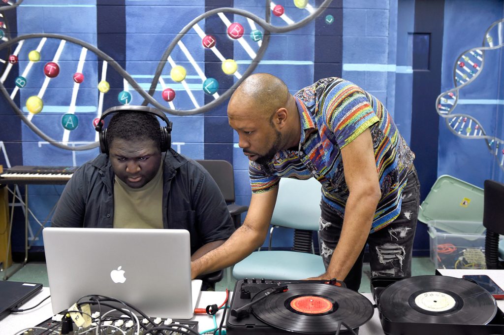 An male instructor leans over a male student wearing headphones at a laptop in the Beat Lab at the Carolina Hip Hop Institute summer 2024.
