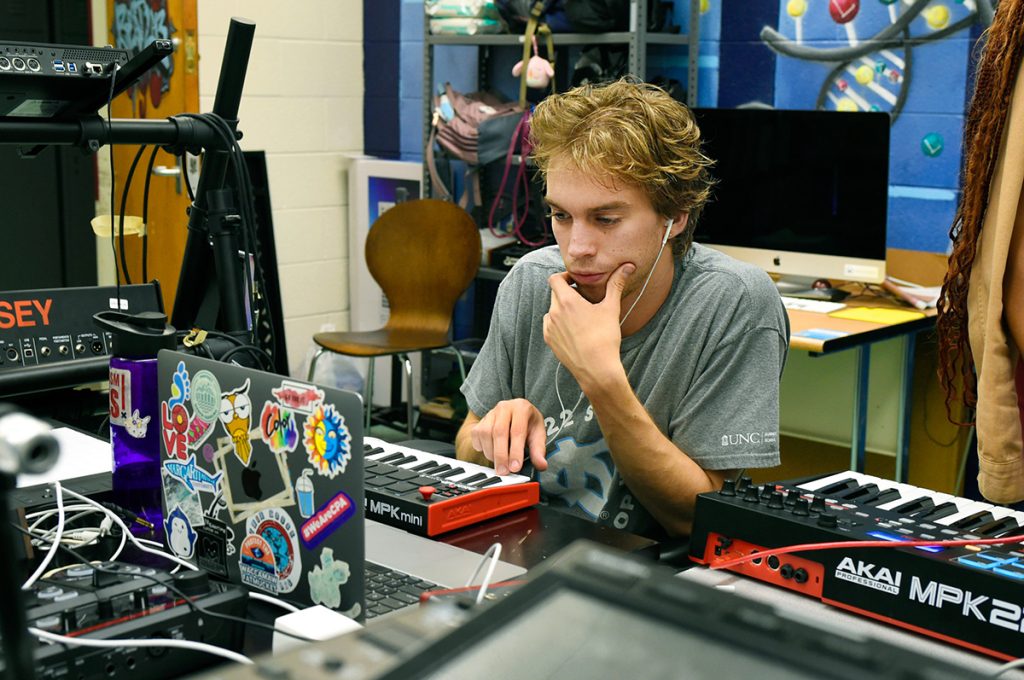 A male student wearing earbuds sits in front of a keyboard in the Beat Lab at the Carolina Hip Hop Institute summer 2024.